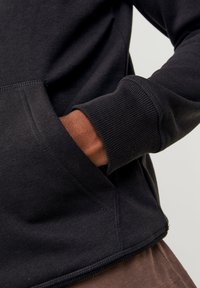 Sudadera negra con bolsillo canguro, puños acanalados y un ajuste holgado, con tejido marrón contrastante visible en la muñeca.