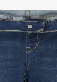 Jeans in denim blu scuro con una cintura blu chiara. Presentano un bottone argentato, cuciture gialle e dettagli nelle tasche frontali.