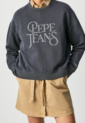 Osoba ubrana w ciemnoszarą bluzę z napisem „PEPE JEANS” wykonanym z białych kropek oraz beżową spódnicę zapinaną na guziki z wiązaniem w talii, z rękami w kieszeniach bluzy.