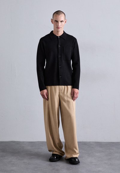 Mann som står innendørs iført en svart skjorte med knapper og brystlomme, beige vide bukser og svarte sko, mot en ensfarget grå vegg.