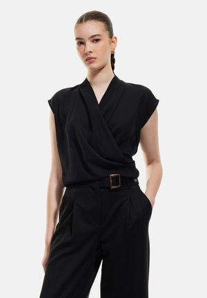 Zwarte wikkelblouse met korte mouwen, zachte textuur en gedrapeerde voorkant. Gecombineerd met hoge zwarte broeken en een riem met gouden gesp.