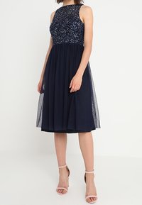 Robe bleu marine avec un corsage perlé et une jupe en tulle. Sans manches, de silhouette évasée, longueur genou, avec une taille cintrée et un voile transparent.
