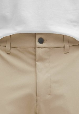 Bež hlače s ravnim prednjim dijelom, šivanim detaljima i metalnim kopčanjem na struku, u kombinaciji s bijelom majicom.