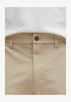 Bež hlače s ravnim prednjim dijelom, šivanim detaljima i metalnim kopčanjem na struku, u kombinaciji s bijelom majicom.