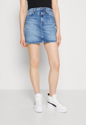 Persoon draagt een blauwe denim minirok, zwart topje en witte hoge sneakers, staand tegen een effen achtergrond.