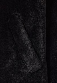 Schwarzer Kunstfellstoff mit einer weichen, plüschigen Textur. Verfügt über eine seitliche Tasche, die von einer glatten Kante umrandet ist, was das einheitliche Erscheinungsbild betont.