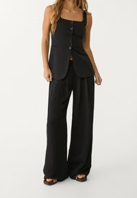 Černý bezrukávový top s rovných střihem, zdobený pěti knoflíky vpředu. V kombinaci s volnými černými kalhotami s širokými nohavicemi.