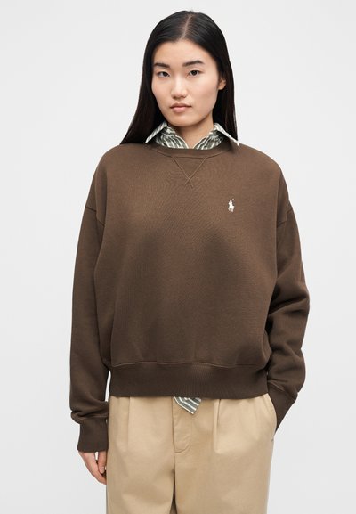 Mlada ženska z dolgimi temnimi lasmi, ki nosi rjavo jopa čez črtasto srajco z ovratnikom in bež hlače, stoji pred belim ozadjem.