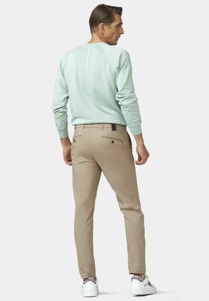 Mann in hellgrünem Langarmshirt, beigen Hosen und weißen Turnschuhen, steht mit dem Rücken zur Kamera und blickt über seine rechte Schulter.