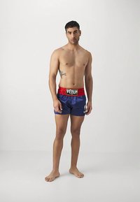 Homem sem camisa, descalço, vestindo calções de boxe Venum azuis e vermelhos, com uma tatuagem no lado direito, em frente a um fundo branco liso.