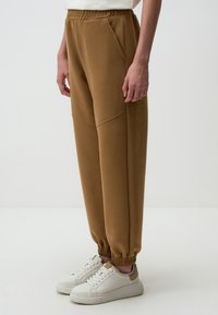 Bruine, taps toelopende joggers met een elastische tailleband en boorden. Voorzien van zijzakken en een gladde stoftextuur voor comfort. G gedragen met witte schoenen.