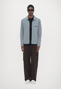 Lys grå zip-jakke med en brystlomme, parret med en sort skjorte og brune cargo-bukser. Model iført hvide sneakers og solbriller.