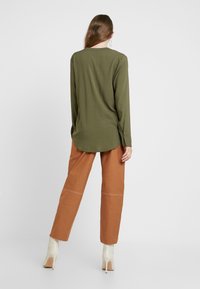 Camicia verde oliva a maniche lunghe con orlo curvo e polsini con bottoni, abbinata a pantaloni color ruggine con dettagli sulle cuciture e stivali alla caviglia color crema.