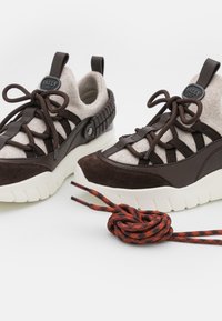 Paire de chaussures de sport marron et crème avec des semelles blanches épaisses et des lacets noués, à côté d'un lacet rouge et noir enroulé sur fond blanc.