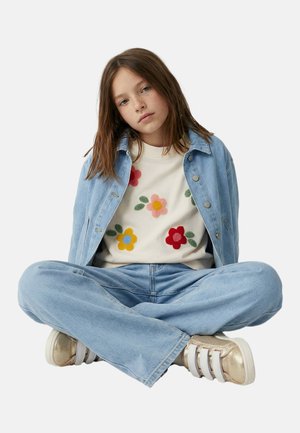 Jeune fille assise en tailleur portant une veste en jean bleu clair et un jean, un pull crème avec des motifs floraux colorés, et des baskets dorées.