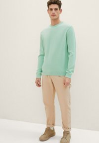 Hellgrüner Langarmpullover aus leichtem Stoff, mit Rundhalsausschnitt. Beigefarbene Cargo-Hose mit seitlichen Taschen und hochgekrempelten Bündchen. Tanfarbene Sneakers.