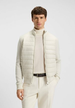 Ljusfärgad puffer väst med hög krage, quiltad design och hel dragkedja. Bärs över en ljus polotröja och kombineras med neutrala byxor.