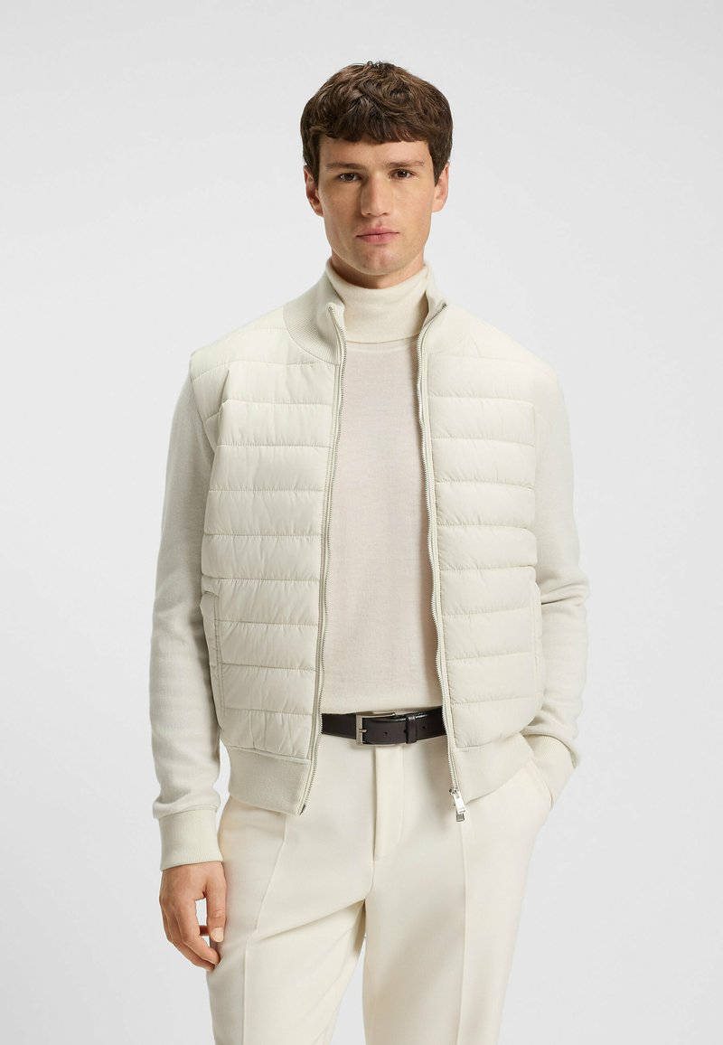 Helle, farbige Steppweste mit hohem Kragen, gestepptem Design und durchgehendem Reißverschluss. Getragen über einem hellen Rollkragenpullover und kombiniert mit neutralen Hosen.