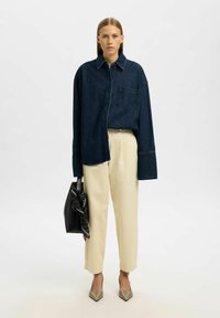 Dunkelblaue Denim-Bluse mit einer Tasche, cremefarbene Hose, schwarze Handtasche mit einem gemusterten Schal und beigefarbene spitze High Heels.