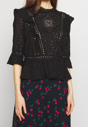 Femme portant un chemisier en dentelle noire à volants et une jupe foncée avec un imprimé cerises rouges, visible du cou jusqu'à mi-cuisse.