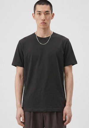Jeune homme aux cheveux courts portant un t-shirt noir uni et un collier chaîne argenté sur un fond neutre.