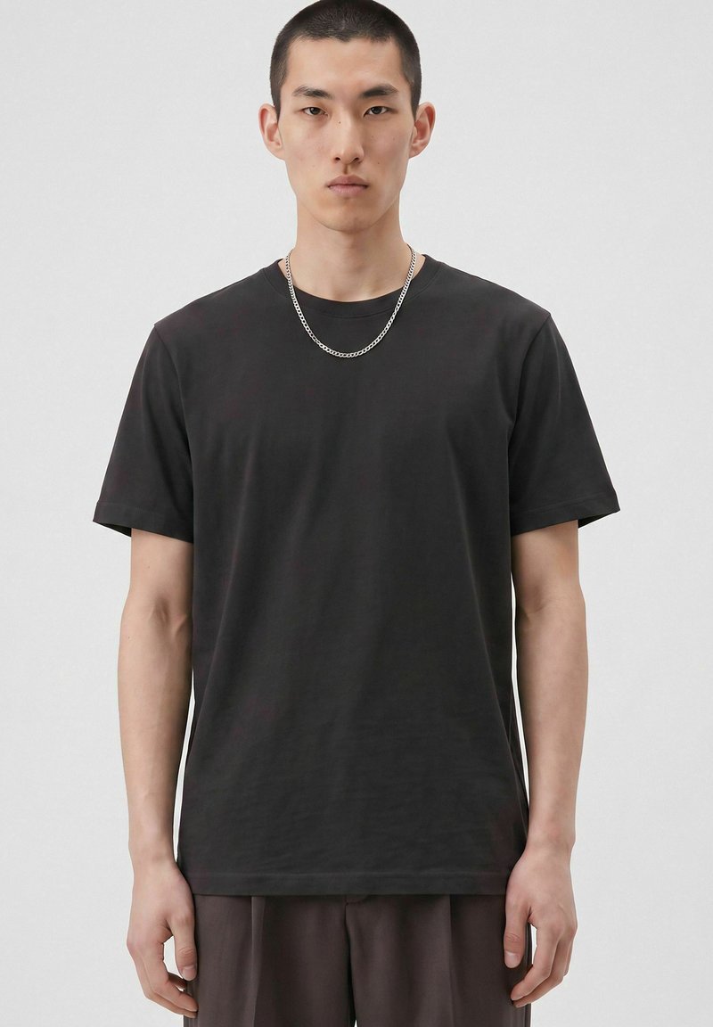 Jeune homme aux cheveux courts portant un t-shirt noir uni et un collier chaîne argenté sur un fond neutre.
