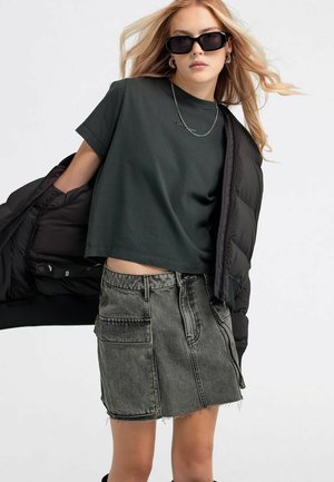 Giovane donna con occhiali da sole neri, t-shirt corta nera, gonna di jeans grigia e giacca piumino nera appoggiata su una spalla.