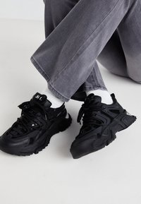 Černé sportovní boty s texturovaným povrchem, síťovanými panely a silnými podrážkami. Šňůrky jsou utažené a viditelné bílé kotníkové ponožky.