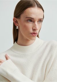 Maglione crema con un alto colletto a costine e polsini a costine. Tessuto strutturato, spalle aderenti e silhouette rilassata. Orecchini argentati visibili.