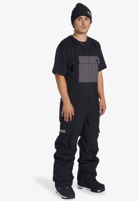 Zwarte overallbroeken met een grijze borstzak, verstelbare banden en cargostijl zakken. Gepaard met een zwart T-shirt en een beanie.