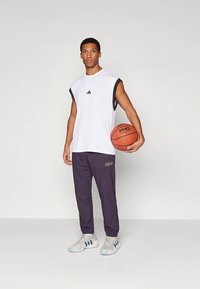 Wit mouwloos atletisch shirt met zwarte accenten, gecombineerd met donkerpaarse broek en grijze sportschoenen. Houdt een basketbal vast.