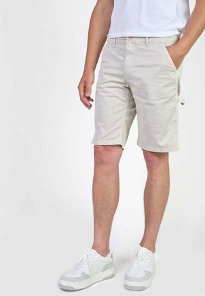 Shorts beige clair en coton, dotés de poches latérales et d'un léger détail cargo. Associés à des baskets blanches et un t-shirt blanc uni.