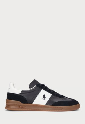 Sort-hvid Polo-sneaker med brun sål, snørelukning foran og Polo-logo på siden og hælen mod en ensfarvet lys baggrund.