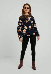 Kvinne som har runde solbriller, blomsterkjole med lange ermer og blonder, svarte leggings og burgunder ankelstøvler, som står mot en grå bakgrunn.