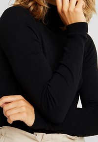 Pull à col roulé noir en tissu maille douce avec manches longues, poignets côtelés et col haut, présentant une silhouette ajustée.