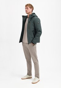 Donkergroene heren hoodie met een slim fit, voorzien van een ritssluiting aan de voorkant, twee zijzakken en een gestructureerde afwerking, gecombineerd met beige broek en witte sneakers.