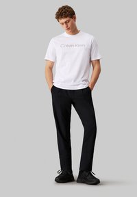 T-shirt bianco in cotone con logo "Calvin Klein", abbinato a pantaloni larghi neri e scarpe da ginnastica nere. Sfondo neutro.