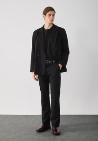 Sacou negru cu două rânduri de nasturi peste un tricou negru, asortat cu pantaloni negri și bocanci burgundy până la gleznă. Textură netedă și design elegant.