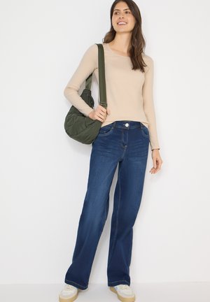 Top a maniche lunghe color beige chiaro abbinato a jeans blu a gamba larga. L'outfit è completato da una borsa a tracolla trapuntata verde e sneakers bianche.