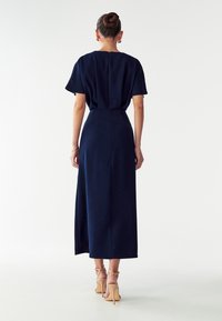 Tummansininen, lyhythihainen maksimekko, jossa on istuva vyötärö ja suora siluetti. Mukana takana oleva vetoketju ja sileä tekstuuri.