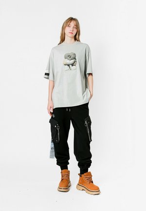 Camiseta oversized gris claro con un gráfico impreso, combinada con pantalones cargo negros con cremalleras y grandes bolsillos, y botas marrones hasta el tobillo.