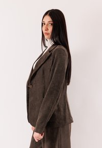 Blazer in velluto a coste marrone con design aderente, chiusura a bottone singolo, rever a lancia e tasche laterali, mostrato di profilo su uno sfondo chiaro.