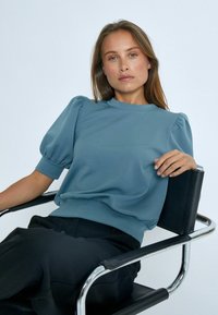 Svetle modrý, krátký rukáv, mikina s balónovými rukávy a kulatým výstřihem, v kombinaci s černými kalhotami, sedící na moderní židli. Hladká textura.
