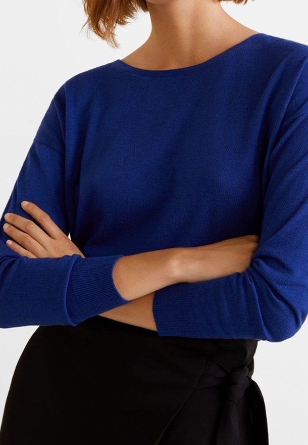Femme portant un pull bleu à manches longues, les bras croisés sur une jupe portefeuille noire nouée à la taille.