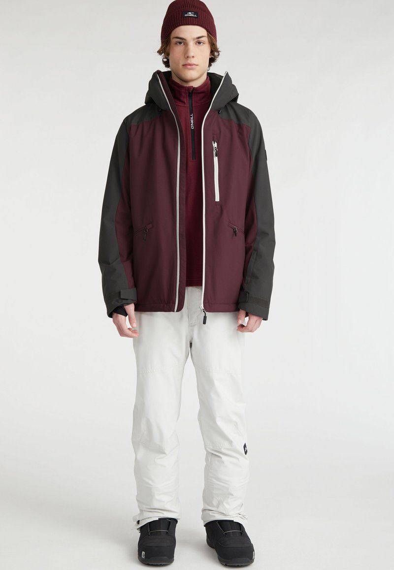 Maronrote und graue wasserfeste Jacke mit Kapuze, schwarzem Reißverschluss und Seitentaschen. Kombiniert mit weißen Hosen und schwarzen Schneestiefeln.