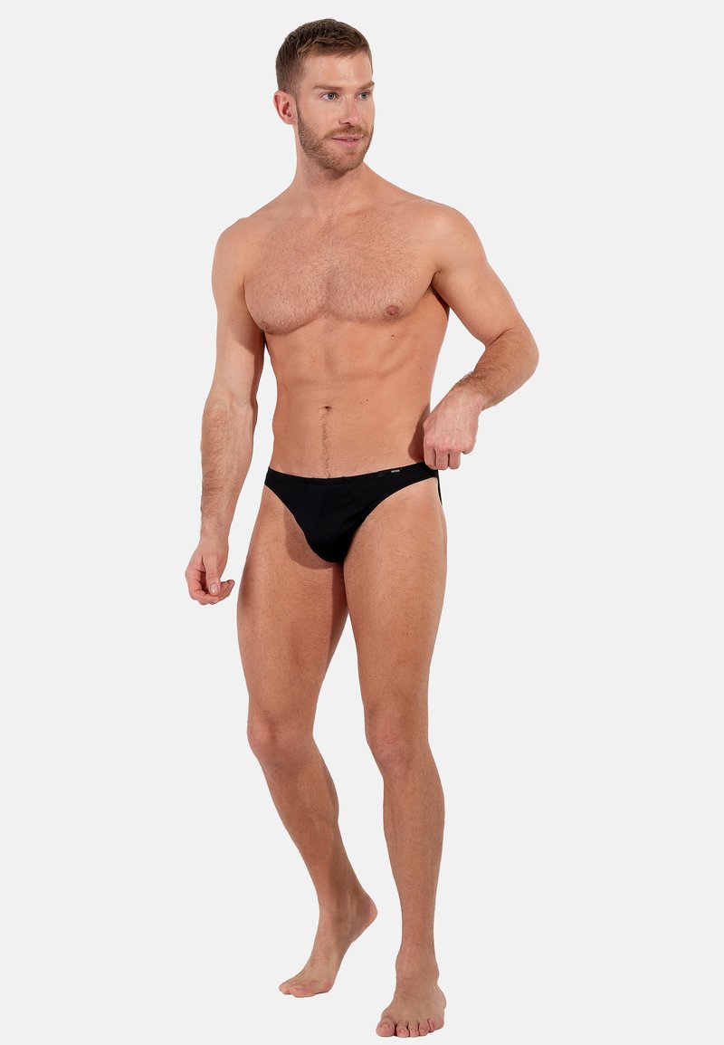 Schwarzer Bikini-Brief aus weichem Stoff, mit einem niedrig geschnittenen Schnitt, elastischem Bund und minimaler Bedeckung. Das Model posiert barfuß stehend.