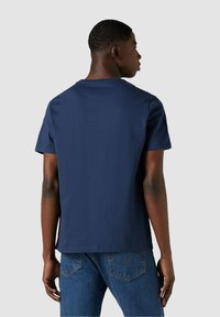 Giovane uomo di spalle vestito con una semplice maglietta blu navy e jeans blu, su uno sfondo grigio chiaro.