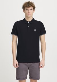 Sort polo-shirt med krave, har en knaplukning og kontrastgrå kant på ærmerne. Lille logo centreret på brystet.
