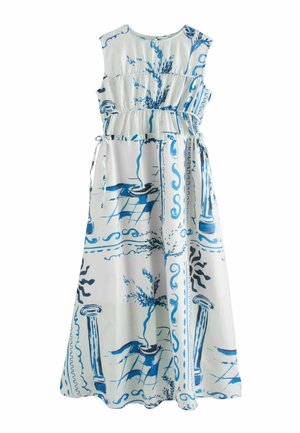 Frau steht auf einem Küstenweg und trägt ein langes beiges Kleid mit blauen architektonischen und botanischen Mustern, sie blickt aufs Meer unter klarem Himmel.