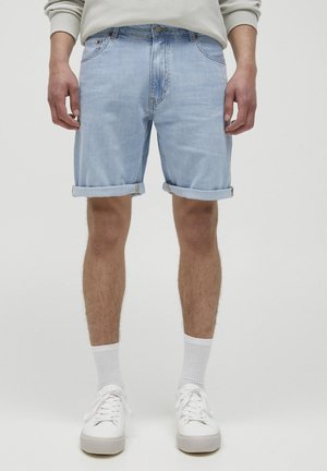 Homme portant un short en jean bleu clair retroussé, des chaussettes blanches et des baskets blanches, debout devant un fond uni.